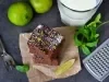Pedaços de bolo de chocolate empilhados sobre papel manteiga, decorados com açúcar de confeiteiro e raspas de limão. Limas inteiras e uma fatia de limão ao lado, com um copo de leite e utensílios, sugerindo um momento de degustação.