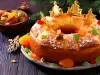 Fotografia de um bolo festivo, decorado com frutas cristalizadas coloridas e coroas douradas, apresentado em um prato prateado.