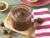 Fotografia em tons de roxo em uma bancada de madeira esverdeada, um pano branco com listras roxas, uma tábua de madeira, um potinho cilíndrico de vidro com o creme de avelã dentro dele. Ao fundo, mini tortinhas com o creme e avelãs espalhadas na mesa.