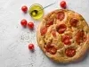 Fotografia vista de cima de uma focaccia com tomate cereja assado, sal grosso e azeite. Ao lado da focaccia tem um recipiente de vidro pequeno com azeite, ao lado alguns tomates cereja, sobre uma mesa branca com texturas.