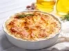 Em cima de uma mesa, refratário com batata gratinada, decorado com cebolinha alecrim.