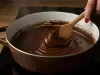 Fotorafia de uma mão feminina segurando uma colher de pau mexendo uma panela grande, branca e marrom, com brigadeiro cremoso de chocolate.