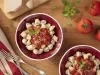 Imagem vista de cima de dois bowls vermelhos com nhoque de mandioca e molho de tomate. Ao lado, tomates, manjericão, garfos e queijo parmesão.