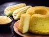 Fotografia de um bolo de milho cortado, revelando seu interior macio, acompanhado de uma tigela de fubá e duas espigas de milho. A cena rústica transmite um apelo caseiro.