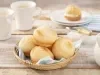 Fotografia em tons de azul em uma bancada de madeira clara, uma cesta de vime com um guardanapo de papel azul e vários pães de queijo dentro dele. Ao lado, um potinho com patê, xícaras brancas e bule de café branco.