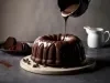 Bolo de chocolate em forma de bundt, colocado em uma tábua clara. Chocolate derretido escorre sobre o bolo, enquanto fatias estão dispostas em um prato preto ao lado. O cenário é elegante, com um jarro branco ao fundo.