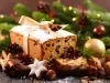 Fotografia de um bolo de frutas decorado com açúcar em pó e um laço, cercado por nozes, anis estrelado e canela, em um ambiente festivo com luzes quentes e ramos de pinheiro.
