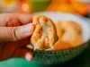 Fotografia de uma mão mostrando metade de uma coxinha aberta de frango e catupiry. Ao fundo tem coxinhas dentro de um recipiente branco e verde fundo.