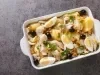 Em cima de uma mesa, refratário com bacalhau, batatas, ovos cortados ao meio e azeitonas pretas, decorado com folhas de manjericão.