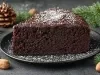 Fotografia de uma fatia de bolo de chocolate escuro em um prato preto, também polvilhada com açúcar de confeiteiro. Elementos como pinhas e nozes ao redor evocam um ambiente rústico e festivo.