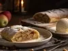 Fotografia em tons escuros de um prato raso de vidro sobre uma mesa de madeira escura. Sobre esse prato tem uma porção do strudel de maçã, ao lado tem uma bola de sorvete de creme generosa em um prato pequeno de vidro.