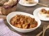 Fotografia em tons de marrom em uma bancada de madeira escura, um paninho roxo listrado, um recipiente branco com a moela de frango com molho de tomate dentro. Ao lado, um cesto de vime com pães, talheres de madeira e um prato menor com uma fatia de pão.
