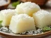 Fotografia de um prato rústico com pedaços de sobremesa de tapioca, cobertos com coco ralado e decorados com folhas de hortelã, em um ambiente tropical, com um fundo que sugere mais ingredientes de coco.