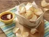 Fotografia em tons de amarelo e verde em uma bancada de madeira, com um pano listrado em amarelo e verde e uma tábua de madeira com uma cestinha de tela com várias batatas chips. Ao lado, um potinho amarelo com ketchup.