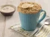 Fotografia em tons de branco e azul de uma bancada de madeira branca com um paninho verde, branco e azul, sobre ele uma caneca azul com bolo de caneca e uma colher. Ao fundo um recipiente redondo com aveia Naturnes.
