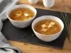 Fotografia em tons de marrom em uma bancada de madeira clara com uma tábua de madeira preta ao centro e dois pratos redondos e fundos com a sopa missoshiro dentro. Ao lado, talos de cebolinha.