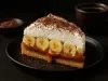 Em cima de uma mesa, prato com fatia de banoffee com massa de biscoitos crocante, doce de leite, bananas em rodelas, decorado com chantilly cremoso e cacau em pó.