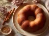 Fotografia de um bolo bundt em um prato cerâmico bege, decorado com flores secas e canela. A composição é elegante, com um pano mauve ao lado.
