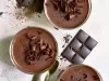 Fotografia vista de cima, em tons claros, de três recipientes de vidro sobre uma mesa clara. Dentro dos potinhos tem a sobremesa, finalizada com ganache e raspas de chocolate, e ao redor tem raspas e algumas barras pequenas de chocolate.