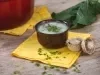 Imagem de um bowl marrom com a receita de caldo verde sobre um pano amarelo. Ao fundo uma panela vermelha e uma folha de couve.