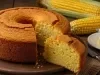 Fotografia de um bolo de milho bem assado, com crosta dourada e interior úmido. Acompanha uma fatia cortada e, ao fundo, espigas de milho frescas e um prato com manteiga.