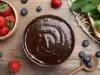 Fotografia vista de cima de um recipiente redondo com a sobremesa, finalizada com ganache, ao lado de um potinho de madeira com morangos inteiros, sobre uma mesa de madeira. Ao redor dos recipientes em mirtilos e mais morangos inteiros, espalhados.