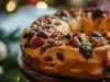 Fotografia de um bolo de frutas tradicional, adornado com frutas secas e nozes, levemente polvilhado com açúcar de confeiteiro, em um ambiente natalino com uma árvore de Natal ao fundo.