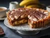 Em cima de uma mesa, banoffee com massa de biscoitos crocante com uma fatia cortada. Dentro da banoffee, bananas em rodelas, doce de leite, decorado com chantilly e canela em pó.
