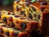 Fotografia de um close de um bolo de frutas, densamente recheado com uvas-passas e outras frutas secas. A superfície brilhante destaca a textura rica, enquanto o interior úmido contrasta com uma cobertura dourada e úmida.