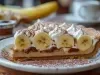 Em cima de uma mesa, fatia de banoffee com massa de biscoitos crocante, doce de leite, bananas em rodelas, decorado com chantilly cremoso e polvilhado com canela em pó.