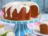 Fotografia de um bolo bundt decorado, coberto com um generoso glacê branco que escorre pelas laterais, adornado com confeitos coloridos. O bolo está em um suporte listrado. Ao lado do suporte, prato branco com uma fatia do bolo fofino e dourado.
