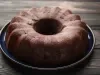 Bolo de chocolate em forma de bundt, com cor marrom escuro, está sobre um prato cerâmico azul. A superfície texturizada sugere umidade, e a apresentação rústica é complementada pela madeira do fundo.