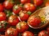 Fotografia de vários tomatinhos cereja confit juntos com bastante azeite e salsinha picada em uma assadeira preta. A imagem mostra uma colher de madeira segurando um dos tomatinhos com azeite.