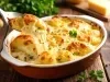Em cima de uma mesa, refratário com batatas cremosas gratinadas com queijo, decorado com salsinha picada. Do lado esquerdo, uma colher pega algumas batatas com queijo derretido.