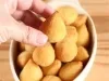 Fotografia de uma mão segurando uma coxinha pequena. Ao fundo tem recipiente branco de vidro oval sobre uma mesa de madeira. Dentro do recipiente tem várias mini coxinhas.