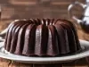Fotografia de um bolo de chocolate em um prato branco decorado, com cobertura brilhante escorrendo pelas laterais. O fundo apresenta uma mesa de madeira rústica e uma jarra de vidro, sugerindo um ambiente aconchegante.