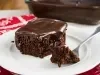 Fotografia de uma fatia de bolo de chocolate em um prato branco, com ganache brilhante e cremosa. Um garfo sugere que a fatia está pronta para ser saboreada.