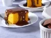 Um pedaço de bolo de cenoura bem amarelinho, servido em um prato branco com um generoso cobertura de chocolate escorrendo pelas laterais. Ao fundo, há outro pedaço semelhante e uma tigelinha com pedaços de chocolate. A textura do bolo parece fofa e leve.