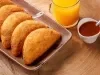 Fotografia de cinco risoles de presunto e queijo sobrepostos em um recipiente retangular marrom escuro sobre uma mesa de madeira. Ao lado dos salgados tem suco de laranja no copo de vidro e um molho vermelho em um recipiente branco pequeno.