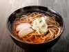 Fotografia mostra um macarrão japonês sendo segurado por hashi, ao fundo um bowl escuro com macarrão dentro.