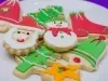 Em cima de uma mesa, prato branco com biscoitos de papai Noel, árvore de Natal e botas, decorados com glacê colorido e confeitos em forma de bolinha.
