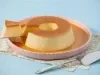 Imagem de um prato rosa claro com pudim de leite condensado com calda de caramelo e uma fatia sobre uma espátula.
