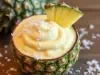 Fotografia em tons claros da metade de um abacaxi com creme homogêneo dentro com coco ralado e um pedaço decorando.