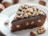 Fatia de bolo de chocolate com recheio cremoso e pedaços de nozes na massa. Cobertura lisa de ganache e finalização com nozes picadas por cima, servida em um prato branco. Um clássico com toque crocante.