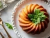 Fotografia de um bolo bundt dourado, decorado com folhas de hortelã, em um prato branco. A composição inclui talheres dourados e coco ralado ao fundo.