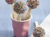 Fotografia em tons de lilás e azul, com copo lilás contendo três pirulitos de brigadeiro com confeitos coloridos, com mais um pirulito ao lado sobre guardanapo colorido, tudo sobre bancada em tons de azul.