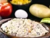 Fotografia de uma salada mista em uma tigela de madeira, com milho, cenoura, uva-passa e um toque de azeitonas, apresentando uma textura cremosa. Ao fundo, um tomate e uma cebola sugerem ingredientes adicionais.