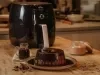 Imagem de um bolo de chocolate com calda de chocolate em um prato. Ao fundo, uma air fryer e ao lado uma jarra pequena com calda.