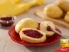 Pãezinhos abertos com geleia vermelha sobre prato vermelho, faca ao lado e mais pães ao fundo, em mesa clara.