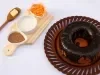 Um bolo de cenoura inteiro, com cobertura de chocolate brilhante cobrindo toda a superfície. Ao lado, há uma tábua com ingredientes usados na receita: manteiga, farinha, cacau em pó e cenoura ralada.